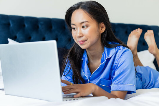 Lying Asian woman wearing blue pajamas typing on laptop in bedroom with tufted navy headboard