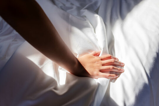 Asian woman pressing bare forearm and hand into sheets in bed, with sunlight creating shadows