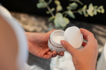 Lifting lid Asian woman holding clear glass cream jar on marble vanity, with eucalyptus sprigs