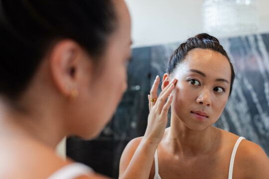 Standing Asian woman touching cheek at marble bathroom vanity, with mirror and fixture