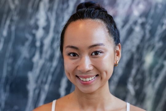 Smiling Asian woman standing in studio before grey marble wall, wearing white tank top, gold hoops