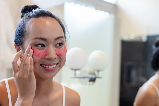Applying hydrogel patches under eyes Chinese woman at bathroom mirror, with globe lamps, copy space