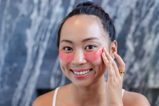 Applying pink under-eye gel patches Asian woman at marble wall vanity, highlighting gold ring