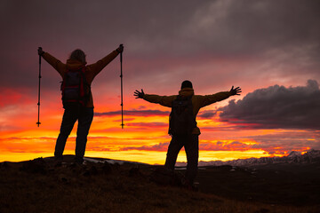 Fototapeta premium Silhouettes of two tourists standing on top of a mountain with their hands raised against a colorful sunset