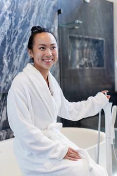 Smiling Asian woman wearing white bathrobe sitting on bathtub rim in bathroom, holding shower wand