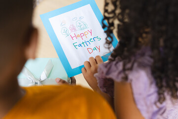 Sitting two siblings holding Happy Fathers Day card at home table, with mint-green gift box