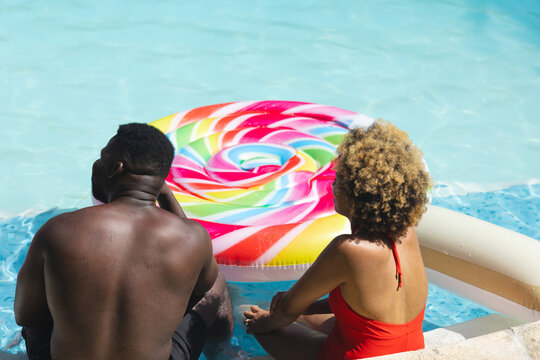 Sitting diverse couple dipping feet in pool at resort poolside, with rainbow swirl float