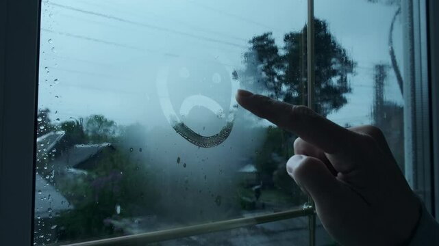 Depressed Mood Concept. Finger Draws Sad Face on Steamy Window While It Rains Outside. Drawing Sad Emotion on Glass. Mood of Isolation and Melancholy. Seasonal depression