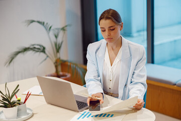 A focused young woman meticulously examines various charts while using her laptop in a stylish office environment