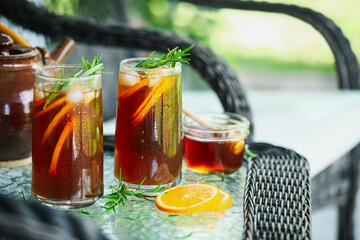 Cold ice herbal tea with orange slices and rosemary stem leaves in  tall glasses. Selective focus on second glass with blurred foreground and background..
