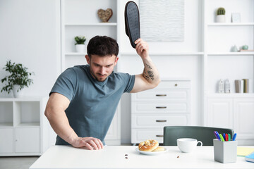 Emotional young man killing cockroaches with slipper at white table indoors. Pest problem