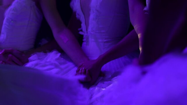Close up of ballet dancers in white tutus holding hands backstage or on stage, captured in soft blue light, showing unity and emotional connection.