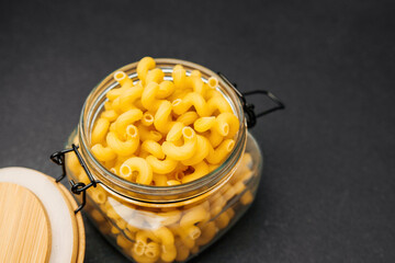 Dried macaroni pasta in a glass jar with wooden lid on black surface