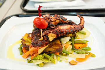 Grilled octopus tentacle served on a bed of mixed vegetables and roasted potatoes with a cherry tomato garnish.