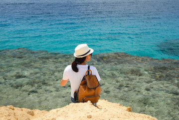 Lonely female traveler in a hat and with a backpack, sitting on a rocky cliff overlooking the seascape.