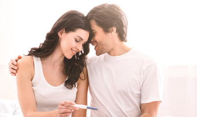 A smiling couple is sitting on a bed at home, with the woman holding a positive pregnancy test. The man has his arm around the woman and they are looking at each other with love and happiness.