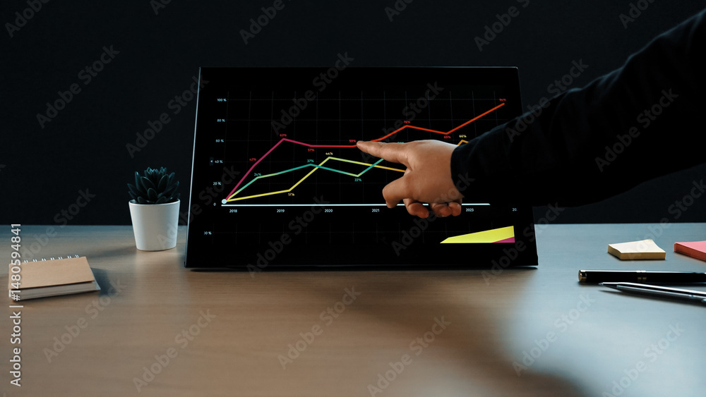 Poster a professional workspace scene featuring a hand pointing at growth trends on a digital graph monitor