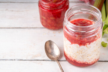 Sweet rhubarb overnight oats in portioned jar. Summer tasty healthy vegan food. Dietary breakfast or snack. Organic rhubarb overnight oats, with rhubarb jam, yogurt, on white wooden table copy space