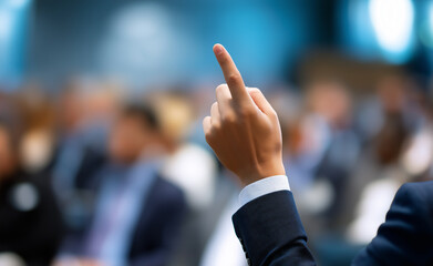 Person in formal attire raising a finger to ask a question in a crowded business conference setting.
