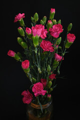 A vibrant bouquet of pink carnations in a glass jar against a black background. The flowers are in various stages of bloom, creating a natural and dynamic composition. Ideal for floristry promotions, 