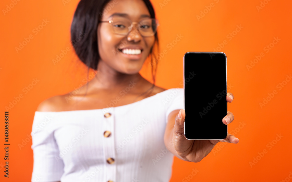 Poster black girl showing phone empty screen smiling at camera standing over orange studio background. sele