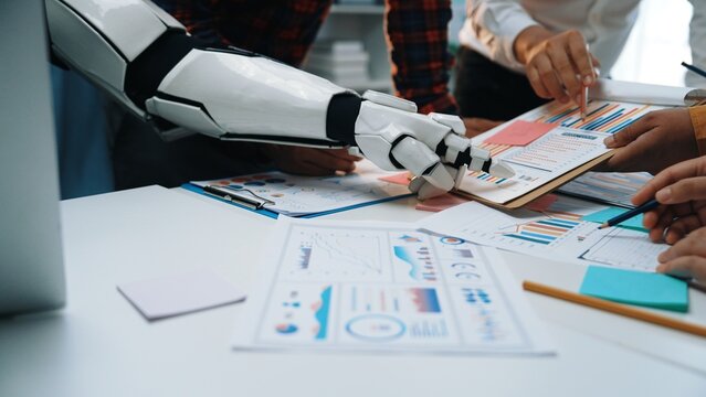 A robot arm collaborates with diverse professionals in a modern office, analyzing reports and charts on a desk, showcasing teamwork and technology integration.Noogenesis