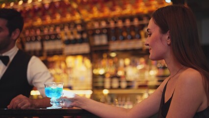 Sitting in smiling beautiful woman guest lively taking with smart bartender while drinking mixed vodka in blue hawaii cocktail and whiskey at luxurious counter bar on Friday meeting at club. Vinosity.