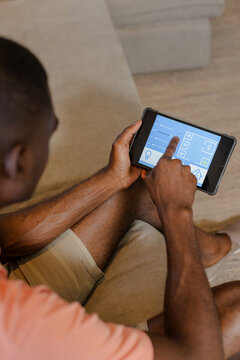 Holding tablet, African American man tapping smart home controls in living room on beige couch