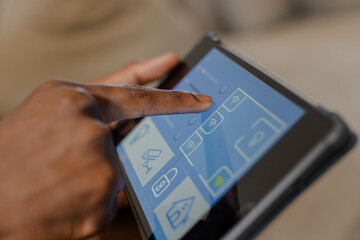 African American man's hand tapping touchscreen tablet in living room, with smart home UI icons