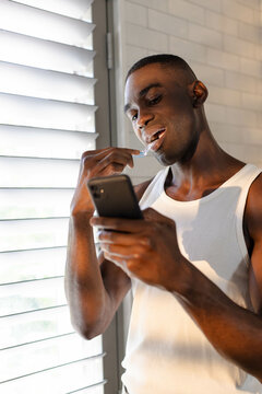 Flossing African American man wearing tank top in bathroom, with smartphone, floss pick and blinds
