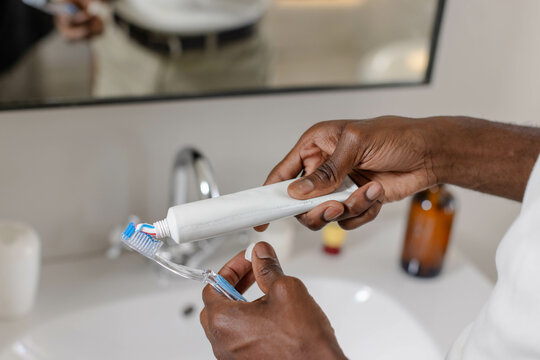 Middle-aged African American man squeezing toothpaste onto toothbrush at sink, with soap dispenser