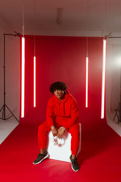 Sitting male wearing red sweatshirt, pants on white cube in studio with red backdrop, headphones