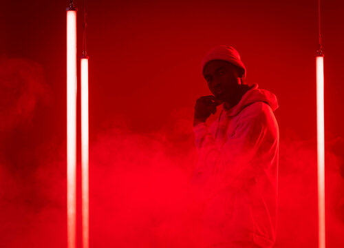 Standing African American man wearing hoodie and beanie amid red smoke and neon tubes in studio