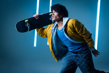 Posing male skateboarder holding skateboard on shoulder in photography studio, with neon lights