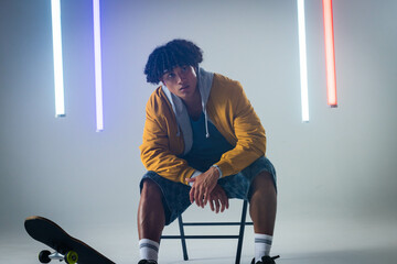 Posing man wearing bomber jacket leaning on folding chair in lighting studio, with skateboard