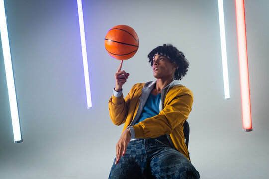 Spinning man wearing mustard jacket on black chair in studio, with basketball, LED tube lights
