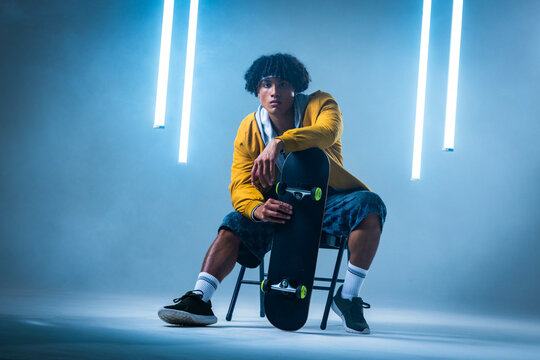 Holding skateboard, African American man in his twenties sitting on chair in studio, with LED tubes