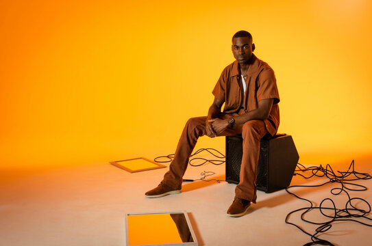 Sitting African American man in brown shirt on speaker cabinet in studio, with cables and panels