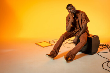 Sitting African American man gazing upward in studio, with amp, cables, lighting panel, copy space