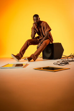 African American man sitting pondering in orange studio, with speaker cabinet cables and panels