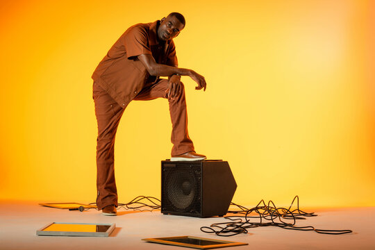 Posing African American man on speaker cabinet in studio, with mic, cables and screens, copy space