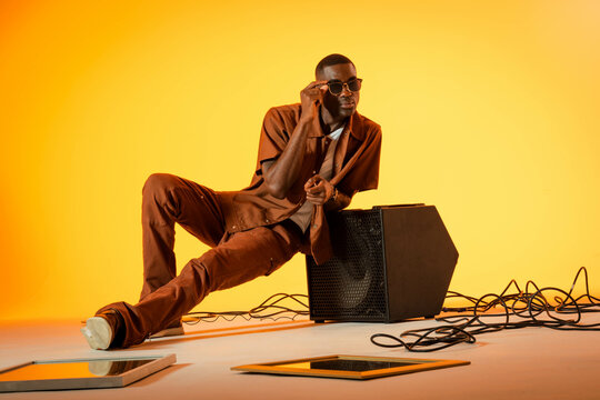 Leaning African American man in brown outfit adjusting sunglasses in studio, with speaker cabinet