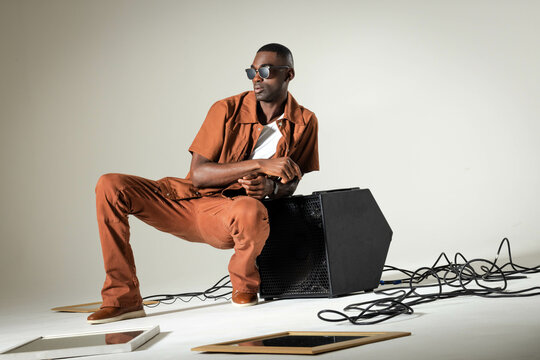 Crouching African American man wearing rust shirt and pants in studio, with speaker, cables, frames