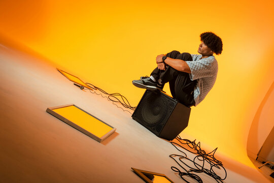 Sitting African American male hugging knees on speaker box in studio, with LED panels, copy space