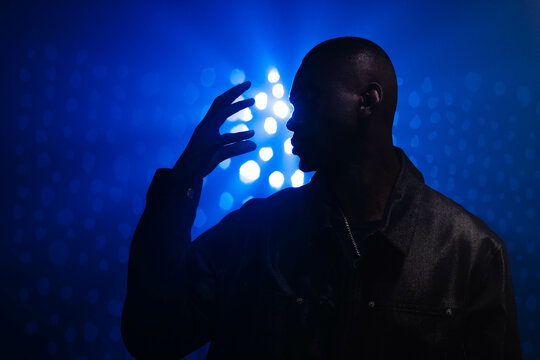 Standing African American man raising hand near face against dark stage, with necklace, blue lights