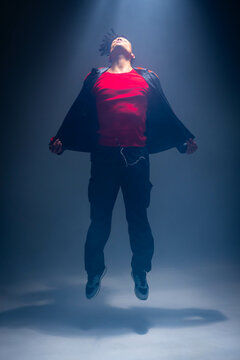 Levitating male dancer wearing red shirt and black jacket in misty studio, under spotlight