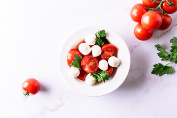 Juicy caprese salad with baby mozzarella cheese, cherry tomatoes and parsley in white ceramic plate on white marble table top view. Healthy diet food concept.