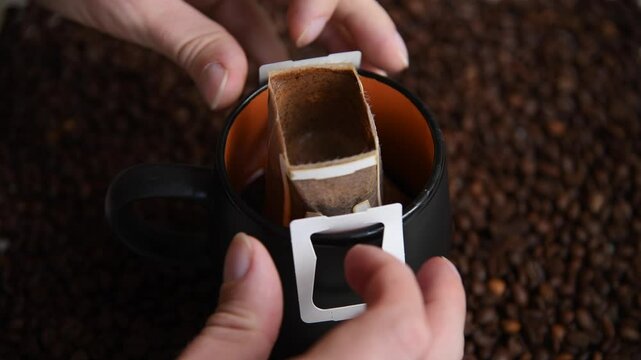 Close-up view of person taking off white paper drip coffee bag from black and orange mug with fresh brewed arabica coffee drink standing on roasted coffee beans.