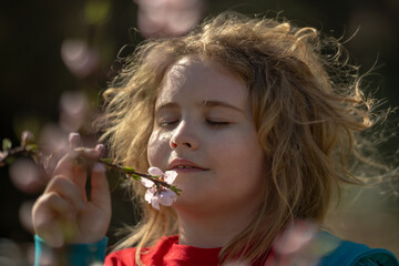 Child face close-up in spring park. Child on spring sunshine. Child smelling flowers spring sunny day. Portrait of kid near blooming tree in March. Child fun in spring. Kid with pink blossom flowers.