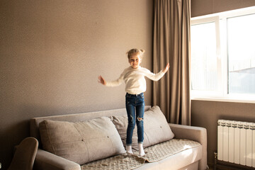 4 years old blonde girl joyfully jumps on couch at home, giggling with excitement. Candid childhood...